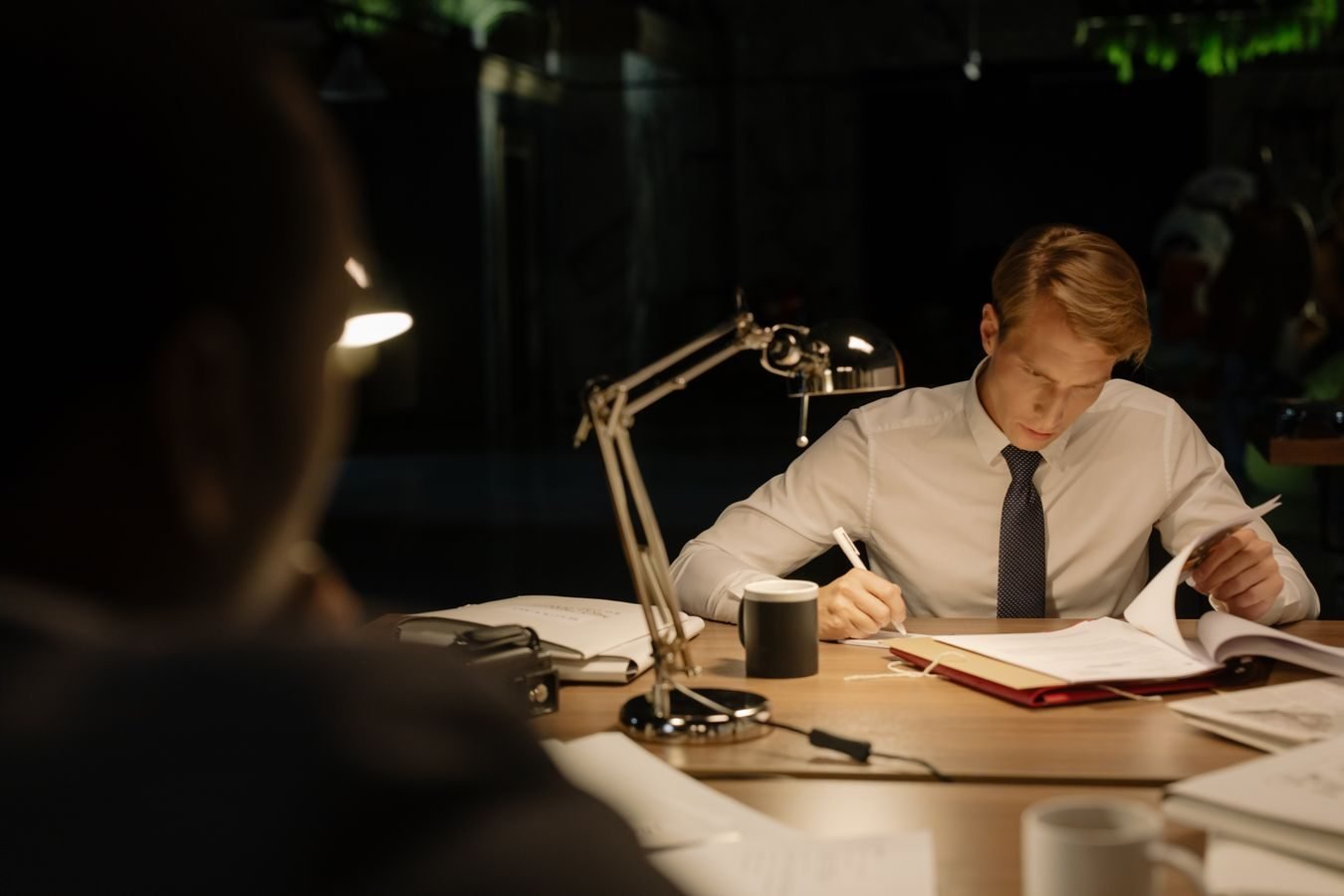 Investigator engrossed in reading documents at a dimly lit desk in an office.
