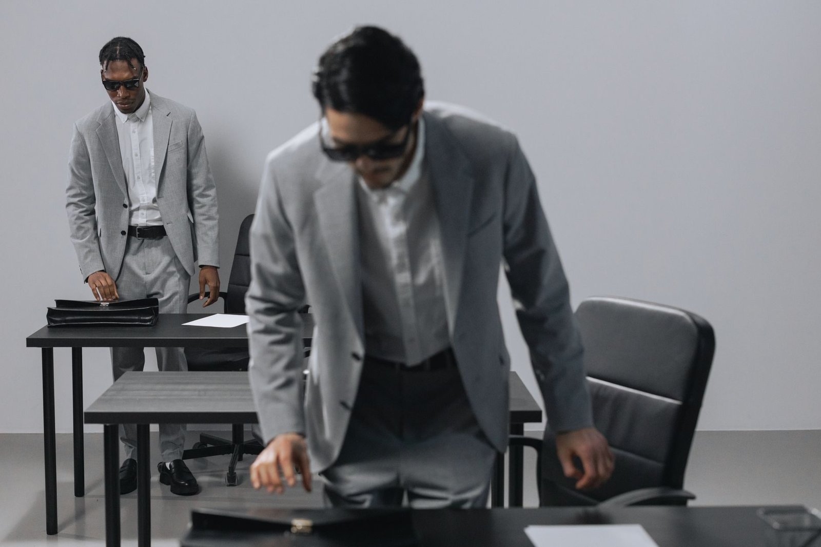 Two businessmen in grey suits and sunglasses in a modern office setting with briefcases.