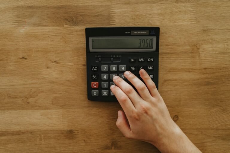 A person's hand pressing keys on a calculator displaying 3750 on a wooden table, top view.