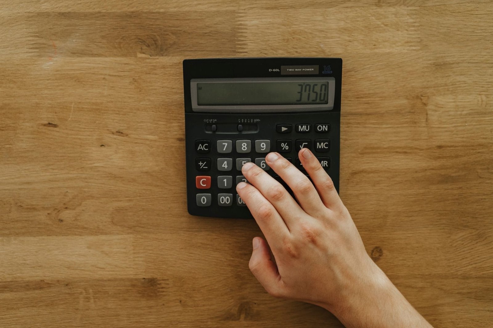 A person's hand pressing keys on a calculator displaying 3750 on a wooden table, top view.