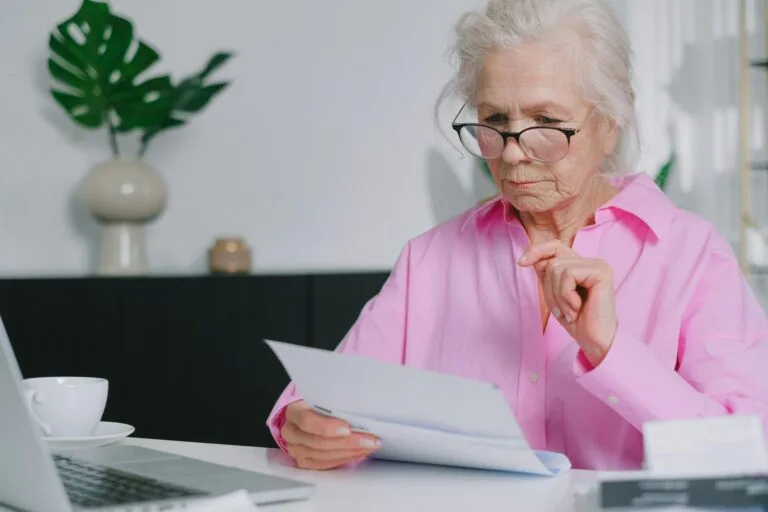 Elderly woman in pink shirt reading documents at home workspace.