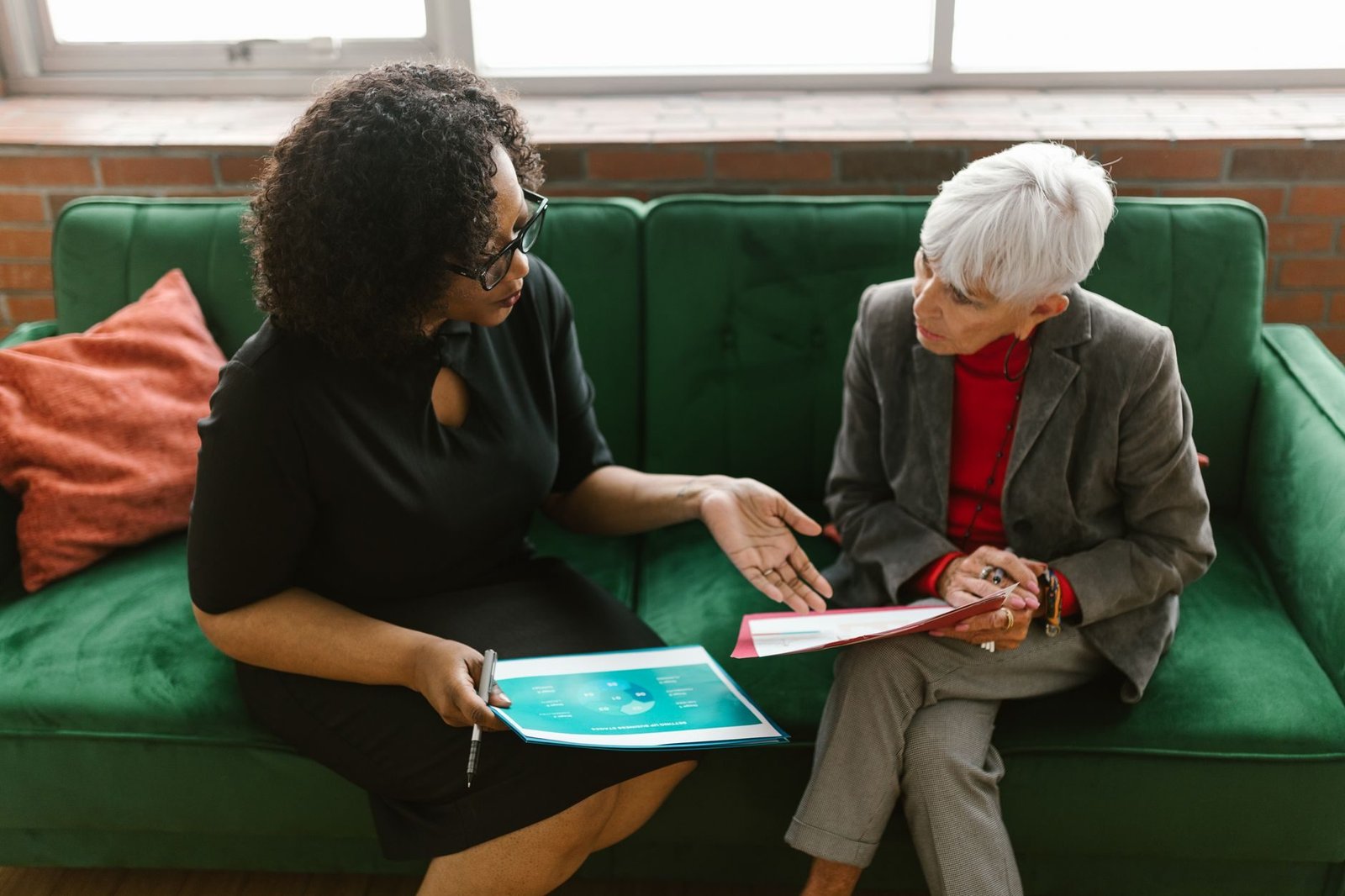 Two women in a business meeting discuss documents while sitting on a green sofa.