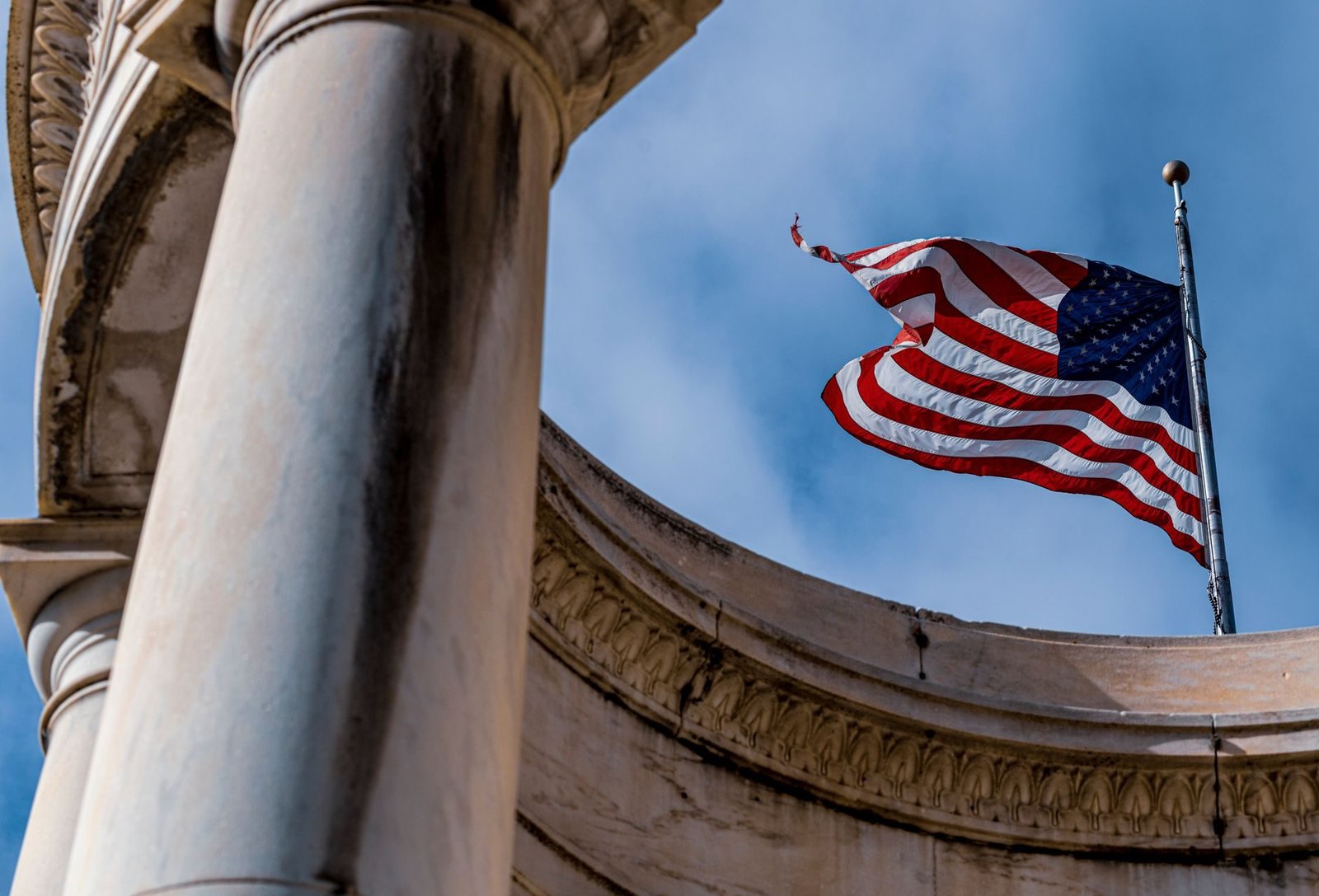 A low-angle view of the American flag waving beside a marble column against a clear blue s
