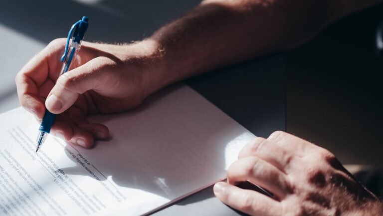 A close-up image of a persons hand signing a document with a blue pen in natural lighting.