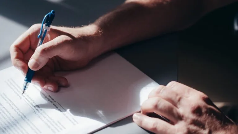 A close-up image of a persons hand signing a document with a blue pen in natural lighting.