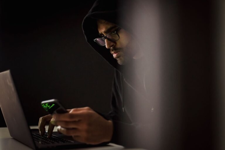 Crop bearded ethnic male computer geek in hoodie chatting on cellphone while typing on netbook in dark room