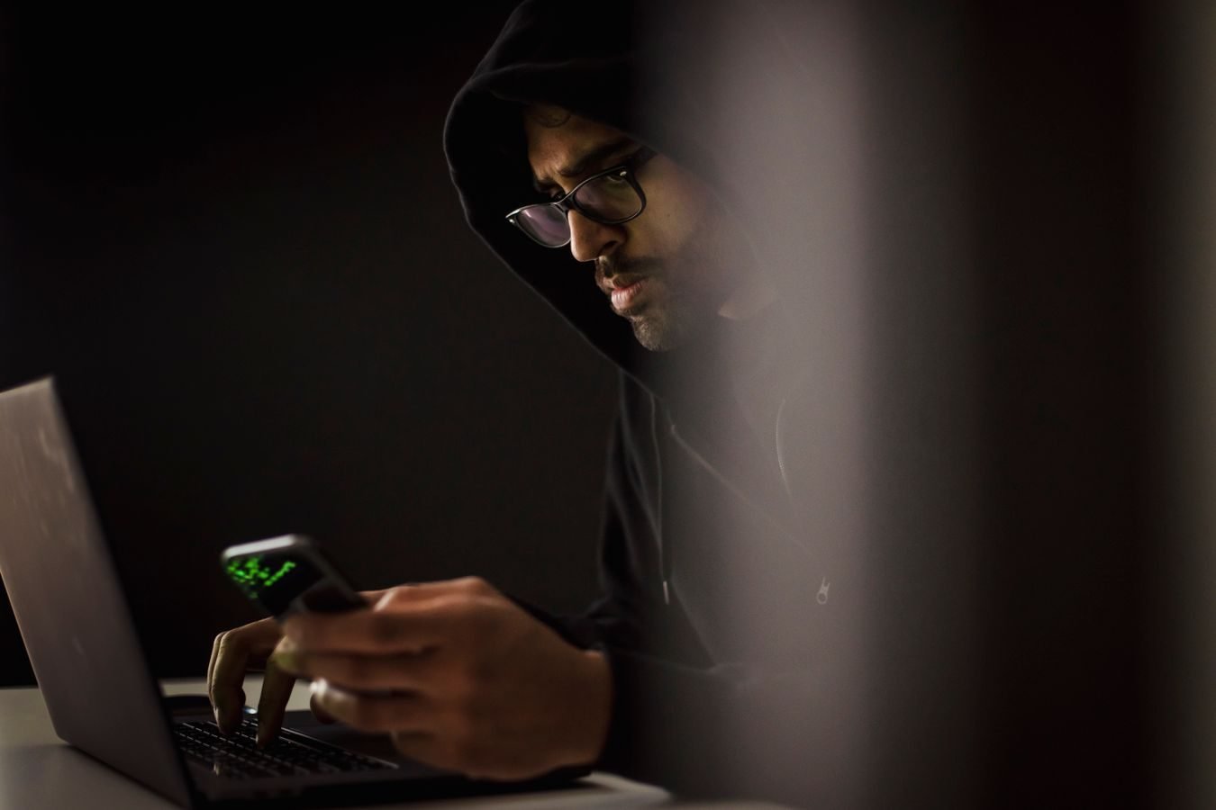 Crop bearded ethnic male computer geek in hoodie chatting on cellphone while typing on netbook in dark room