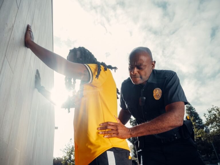 A police officer conducts a frisk search on a suspect wearing a yellow shirt outdoors.