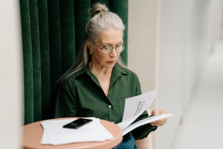 Elderly woman with gray hair reading papers at a small table indoors, focused and thoughtf