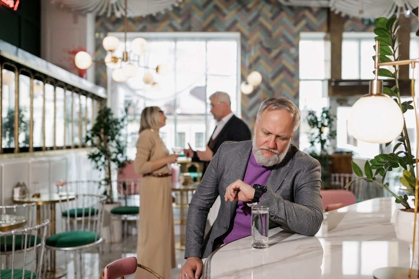 Prazo razoável e silencio administrativo: o que fazer quando o órgão nao responde 1 Elderly man with beard checks wristwatch at stylish cafe counter. Indoor setting with people chatting.
