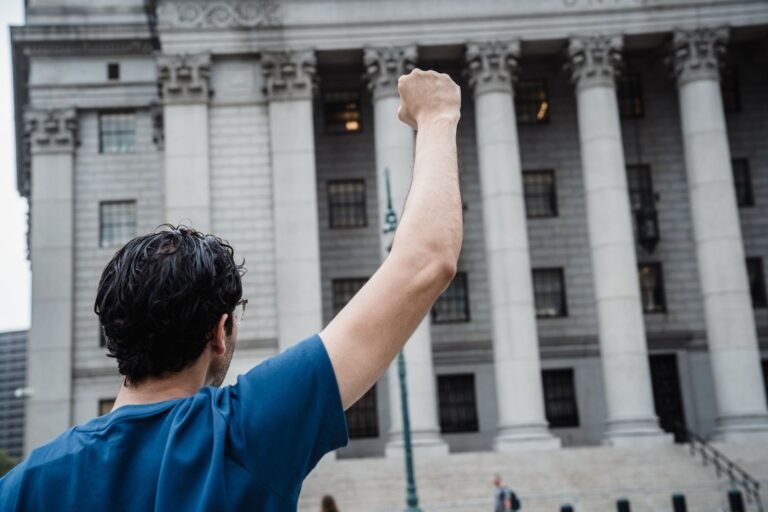A person stands with raised fist in front of a classic architectural building, symbolizing