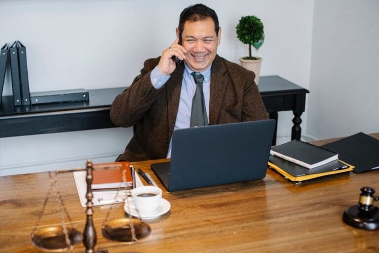 Joyful Asian male lawyer wearing formal suit having conversation on mobile phone and using laptop while working in moder