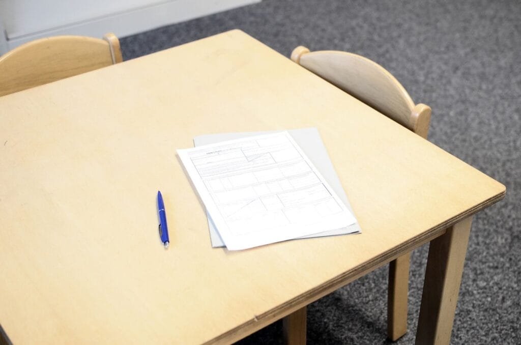 High angle of documents near pen and folder placed on small wooden desk near chairs in light room in school