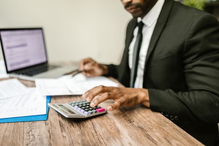 A businessman in a black suit using a calculator at a desk with financial documents and a 