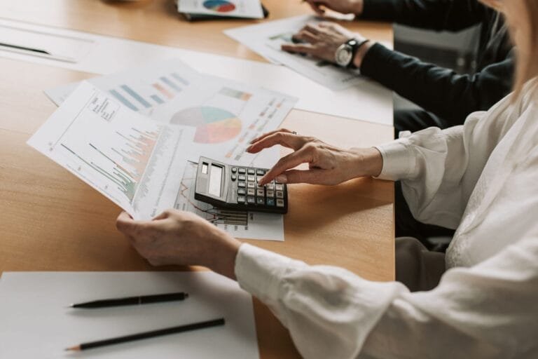 Reforma Tributária 2023: Mudanças e Cronograma 7 Hands performing financial calculations with charts and a calculator at a meeting table.