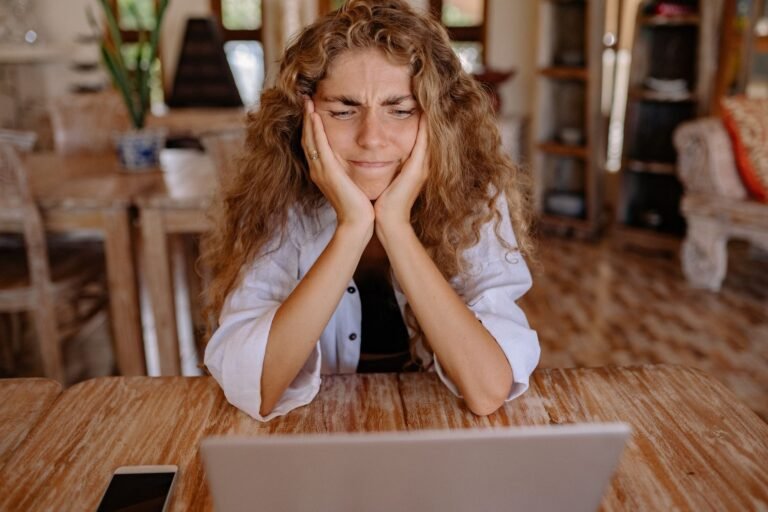 A woman with a frustrated expression works remotely from home using a laptop.