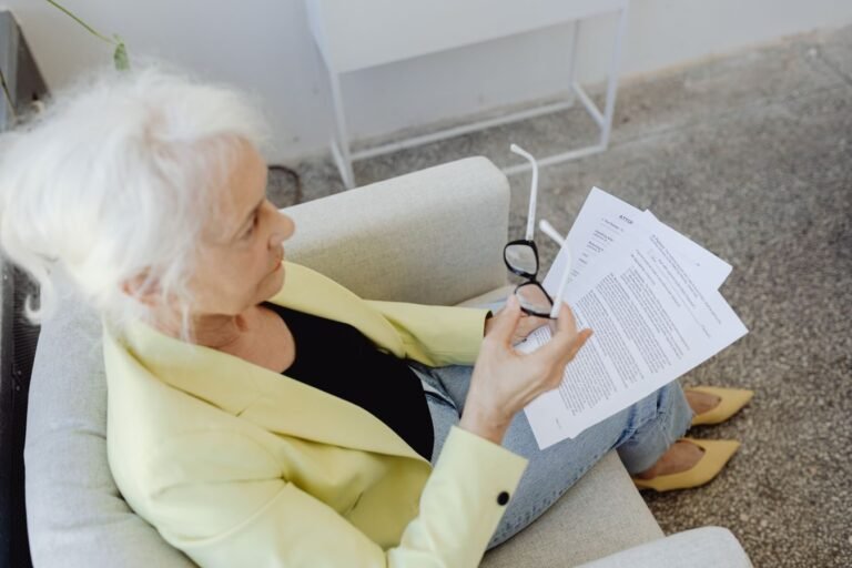 Elderly woman in a blazer holding glasses and documents, sitting indoors.