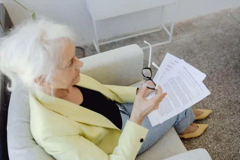 Elderly woman in a blazer holding glasses and documents, sitting indoors.
