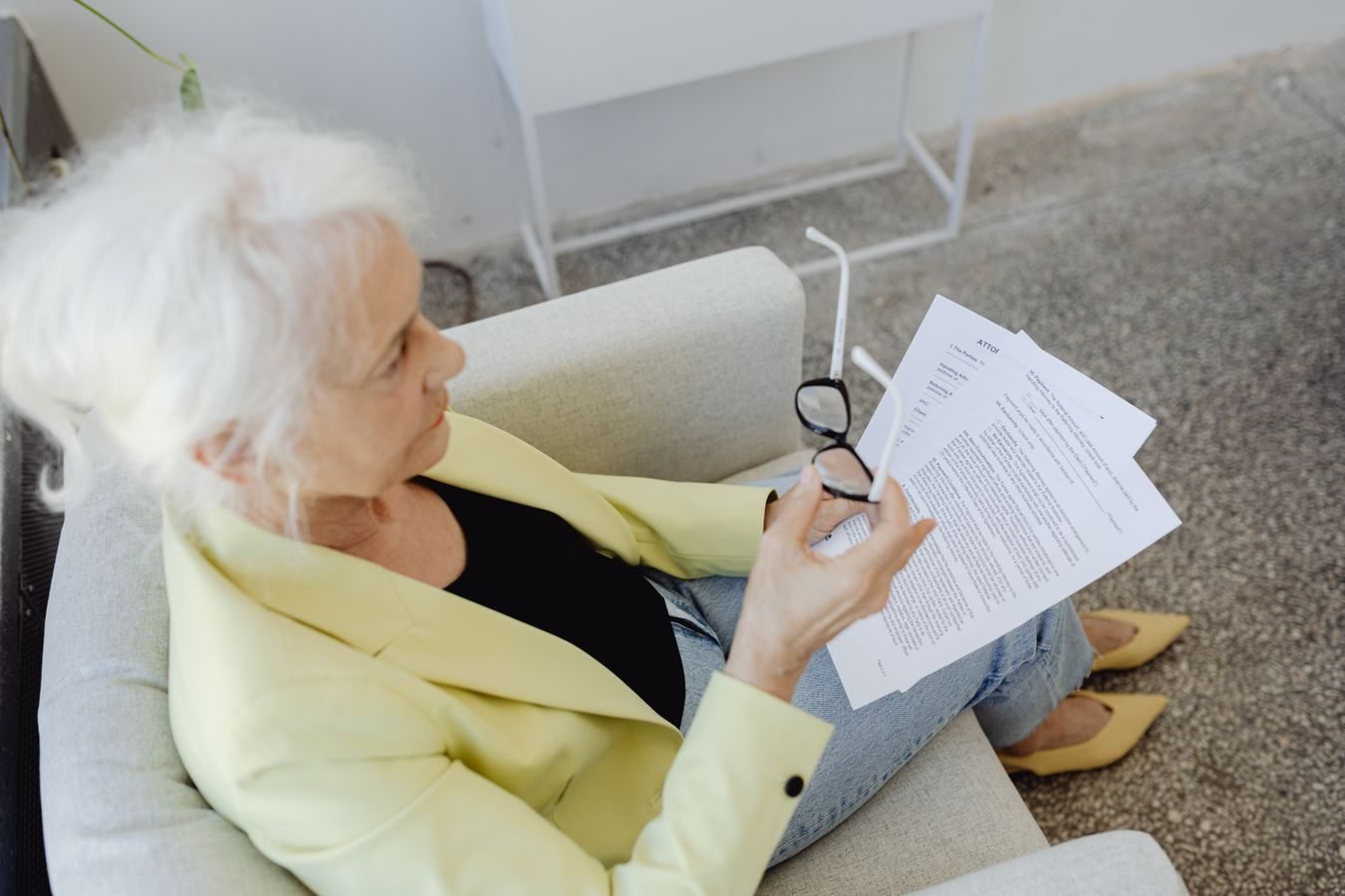 Elderly woman in a blazer holding glasses and documents, sitting indoors.