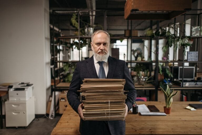Elderly caucasian man with a beard holding piles of paperwork in a modern office environme