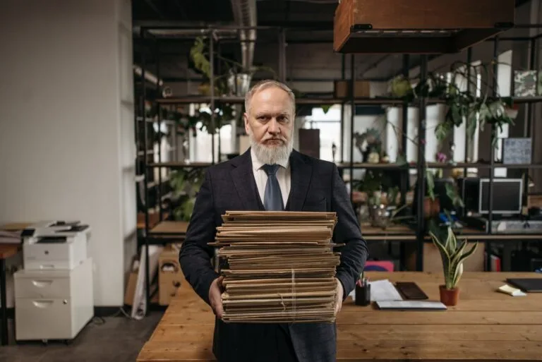 Elderly caucasian man with a beard holding piles of paperwork in a modern office environme