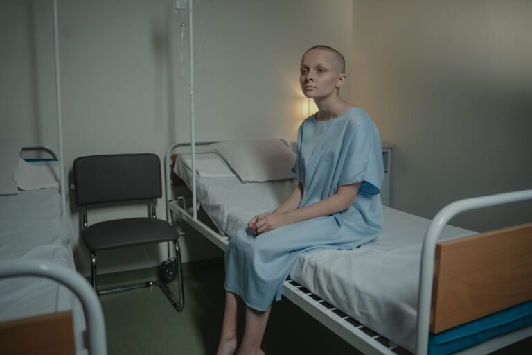 A young woman in a hospital gown sits on a bed in a medical setting, conveying a somber mood.
