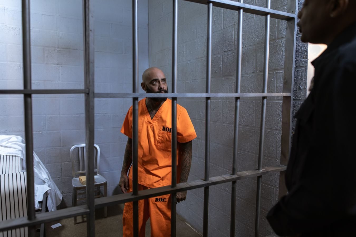 An inmate in an orange jumpsuit stands behind bars with a guard outside the cell.
