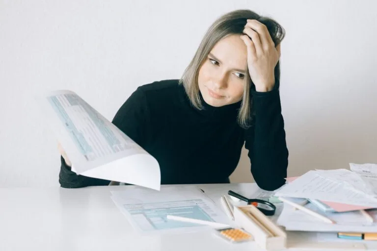 A woman showing stress while reviewing multiple paperwork and financial documents at a des