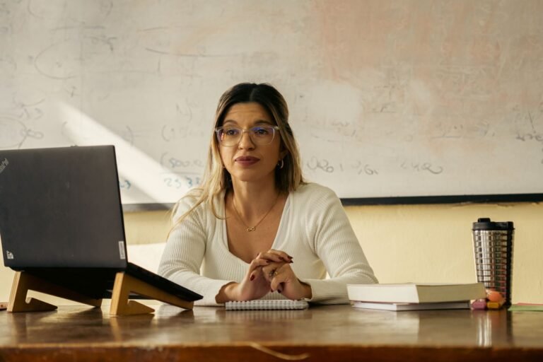 A woman teacher sits at a desk with a laptop and books in a classroom setting.