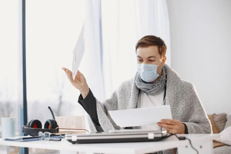 TNU fixa Tema 365 e veda cômputo do período de gozo de auxílio-doença intercalado nas 120 contribuições exigidas para prorrogação do período de graça 2 Man wearing protective mask working from home, wrapped in a blanket, illustrating remote work during illness.