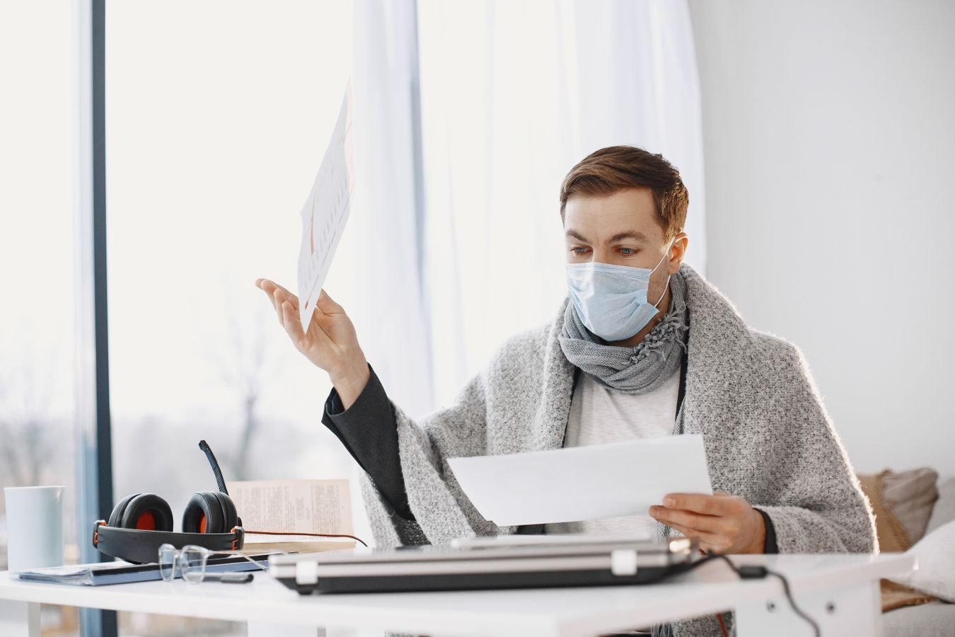 Man wearing protective mask working from home, wrapped in a blanket, illustrating remote work during illness.