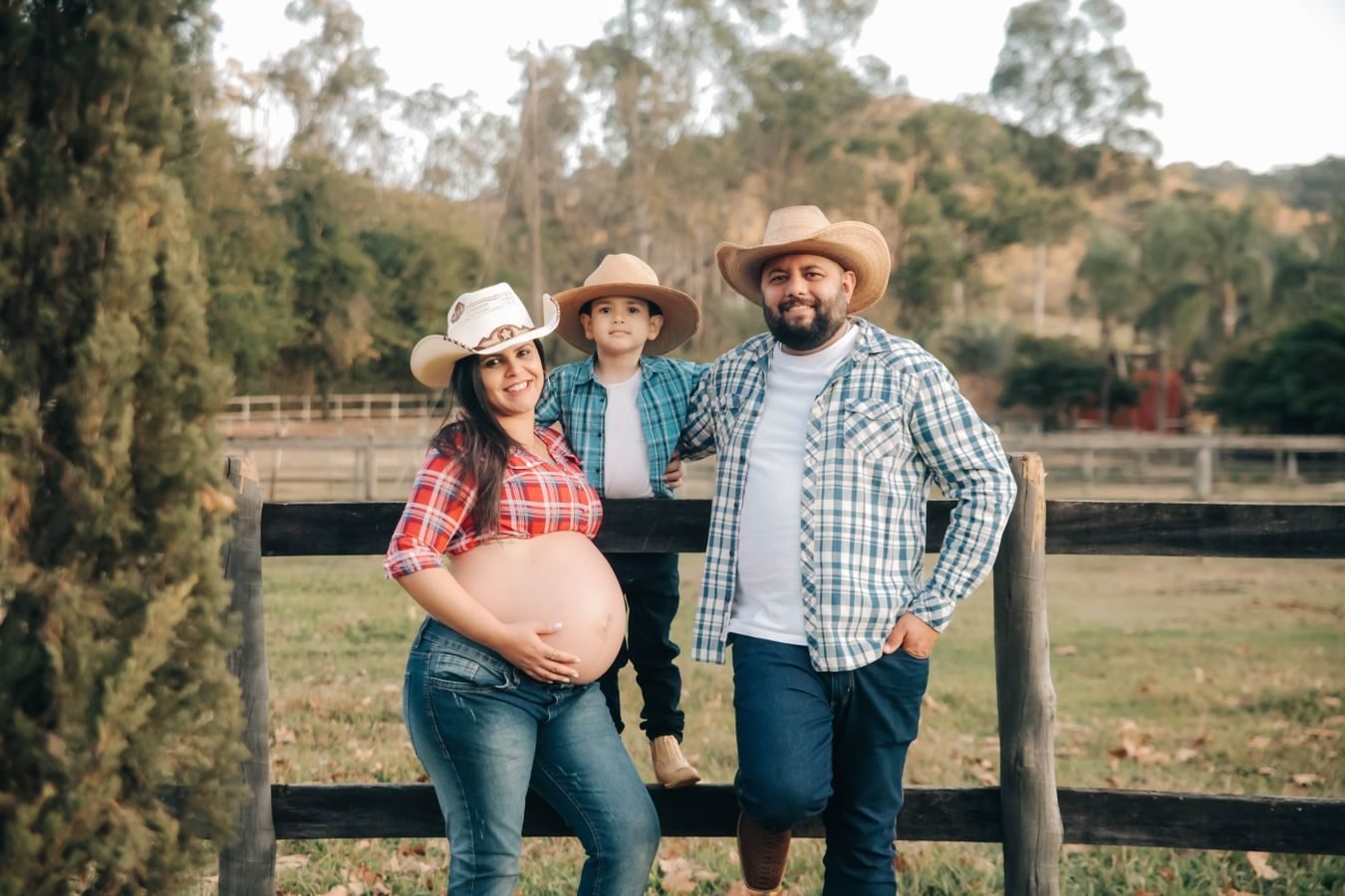 Pregnant woman with family in countryside, smiling and wearing cowboy hats.