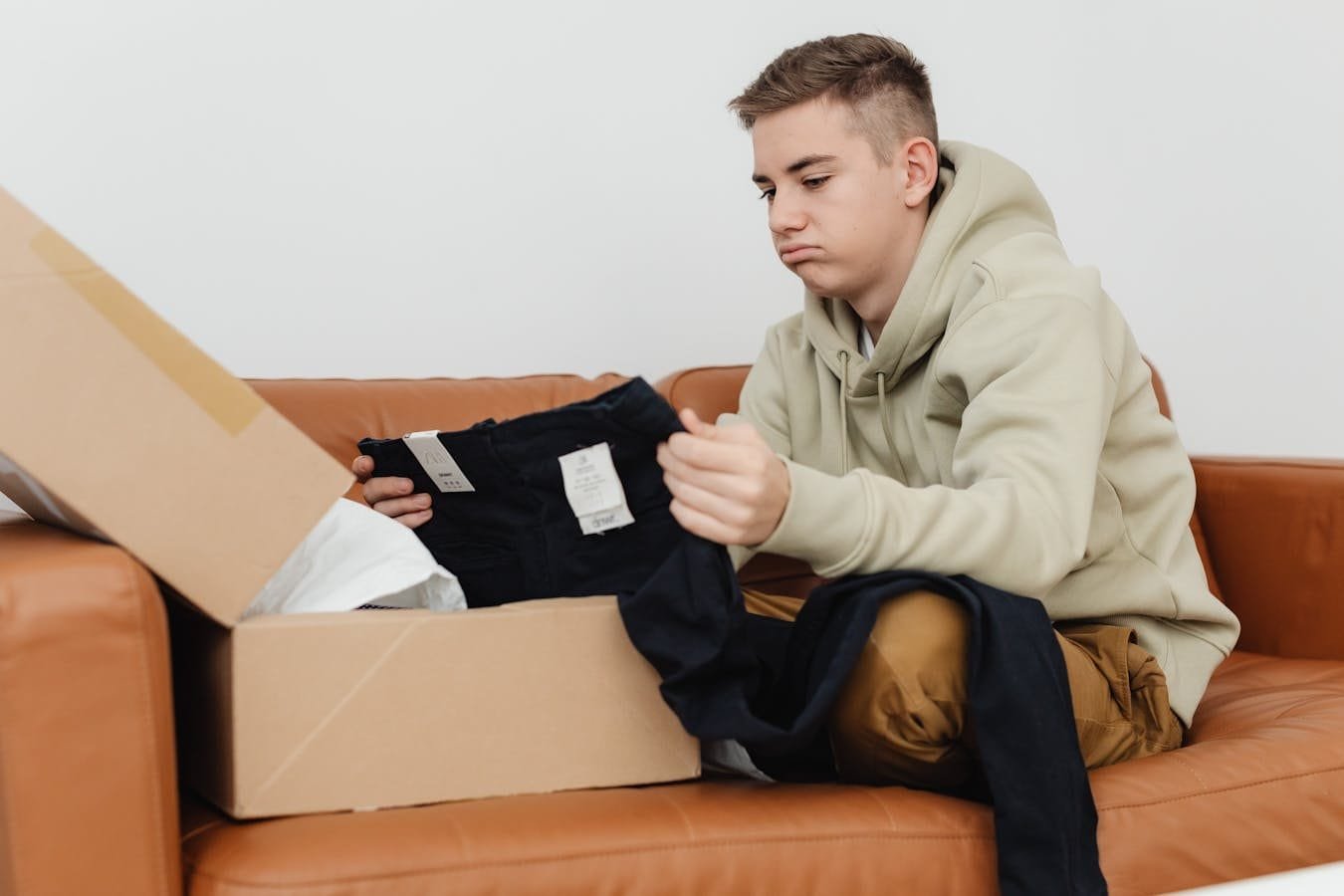 Teenager on couch opening package, unhappy with clothing. Indoors, casual setting.