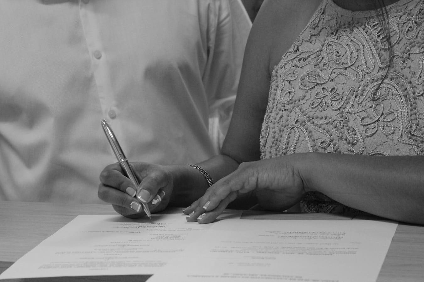 Black and white image of a couple signing documents, focusing on their hands and pen.