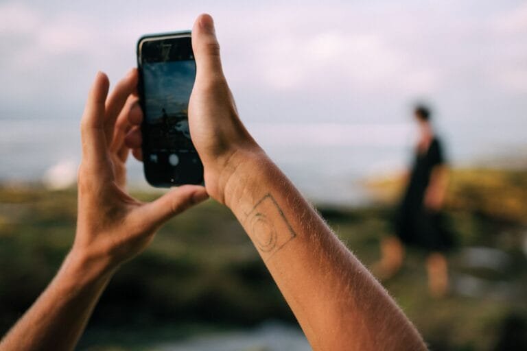 Person taking photo with smartphone at beach capturing another in the distance.