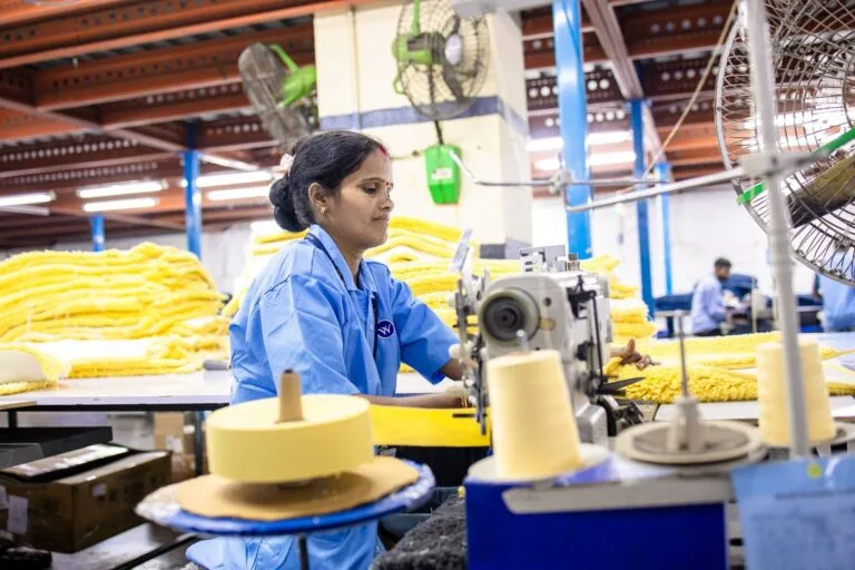 Indian woman working at a textile factory machine with yellow fabric in background.