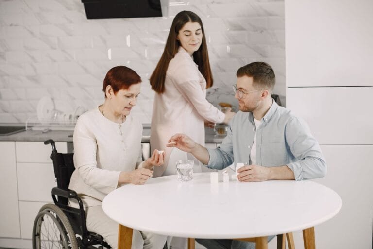 Adicional de 25% por Grande Invalidez: Quem Tem Direito e Como Pedir 2 Elderly woman in wheelchair receiving help with medication from caregiver in modern kitchen.