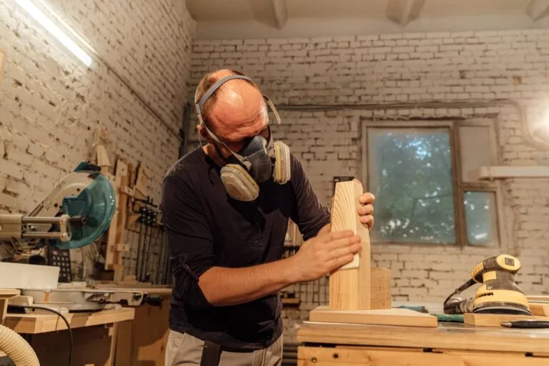 A skilled carpenter wearing a respirator mask sands a wooden piece in his workshop.