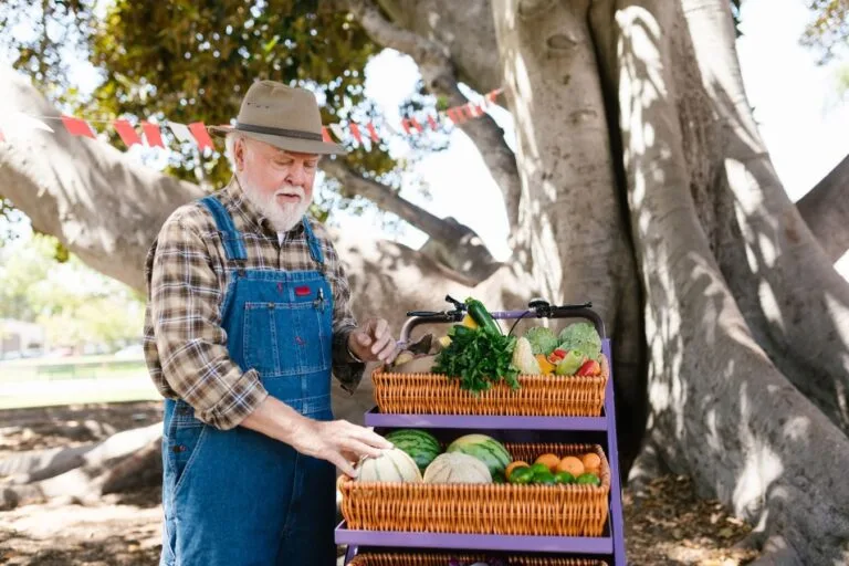 Elderly farmer at a colorful outdoor fruit stand, offering fresh vegetables and fruits.