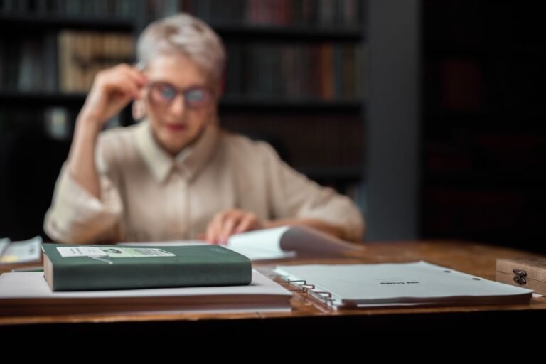 Senior woman in library setting, focused on reading with a book and documents in foregroun