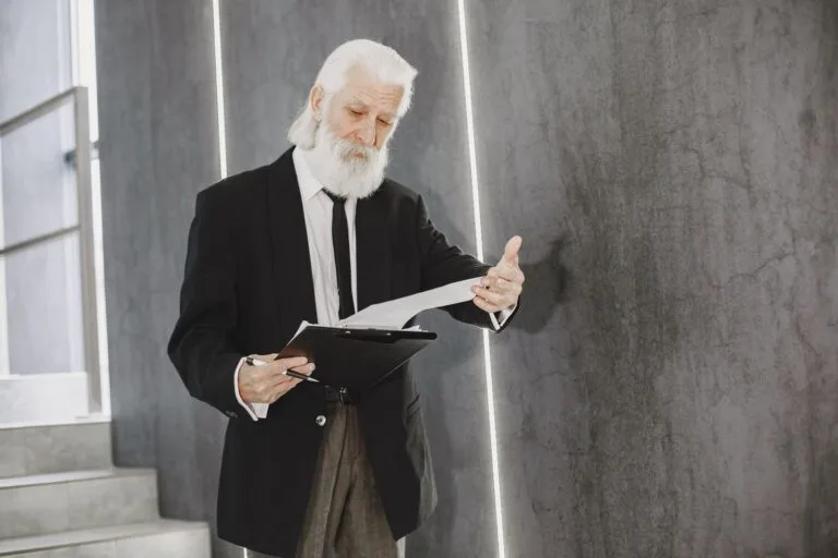 Senior man with white beard in suit reviewing documents indoors, in modern office setting.