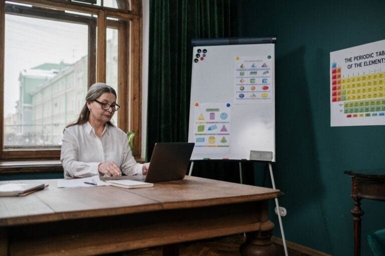 Como podemos ajudar? 7 Elderly woman in classroom using laptop with periodic table and charts.