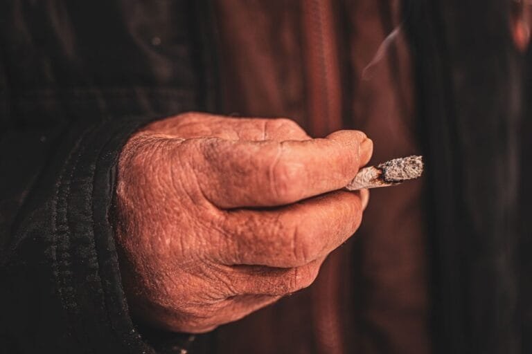 Como podemos ajudar? 11 Close-up of an elderly mans hand holding a cigarette, highlighting skin texture and wrinkles.