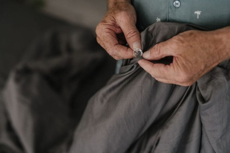 Como podemos ajudar? 10 Close-up of senior hands buttoning a grey textile, emphasizing texture and detail.