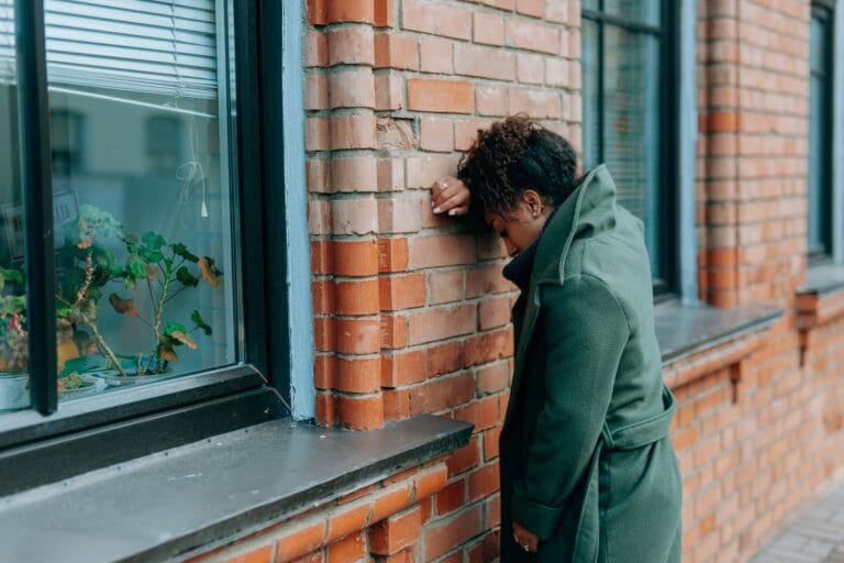 A woman in a green coat leans against a brick wall, reflecting deeply outdoors.