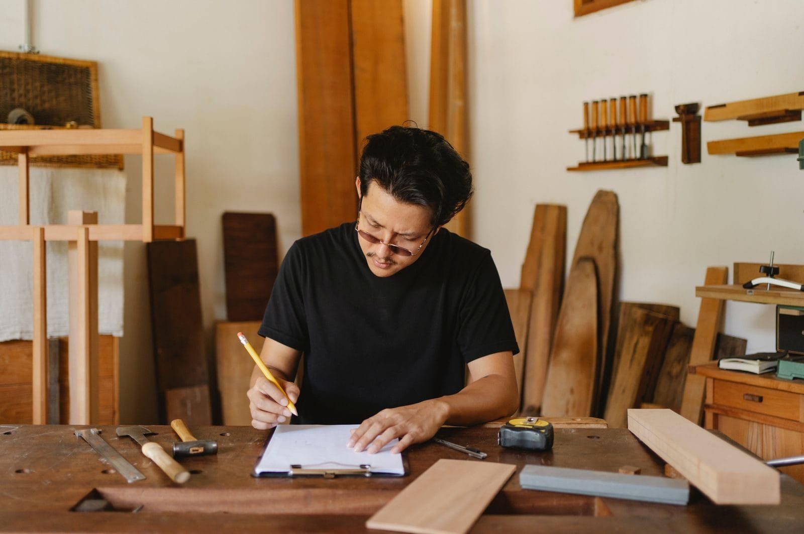 Concentrated ethnic artisan with dark hair in casual clothes sitting at table and writing notes on paper in clipboard wh