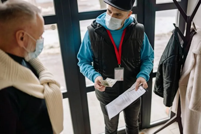 BPC e Bolsa Família: Renda Garantida na Análise 3 A delivery man wearing a facemask receives cash payment from an elderly man indoors.