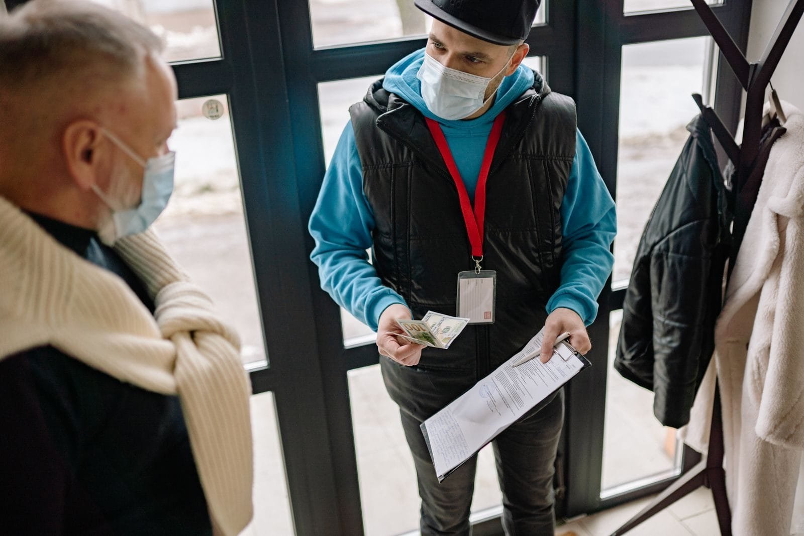 A delivery man wearing a facemask receives cash payment from an elderly man indoors.