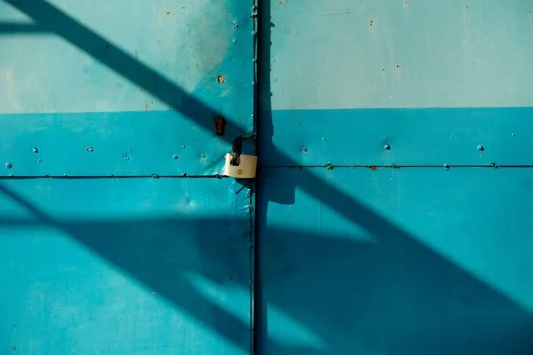 A close-up of a blue metal door secured with a padlock, casting shadows in the sunlight.