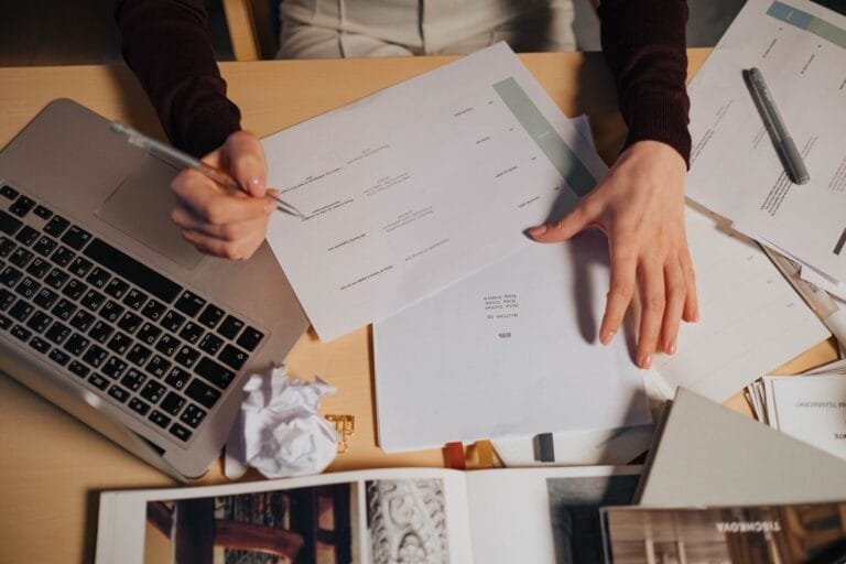 A person taking notes and working on a laptop surrounded by documents on a desk.